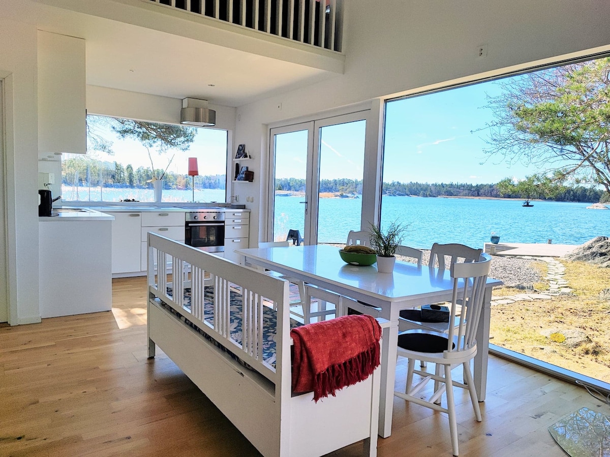 A bright kitchen and dining area is characterized by large windows offering views of the water and surrounding nature. A white table with matching chairs is complemented by a red blanket draped across a bench. The space is open, fostering a relaxed atmosphere.