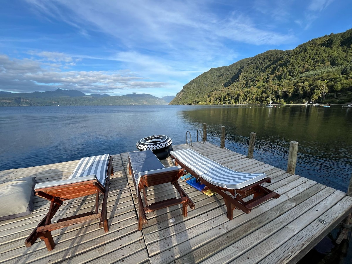 A wooden deck extends over the tranquil waters of Lake Colico, featuring two striped loungers arranged beside a circular inflatable float. Lush green hills rise in the background, with scattered clouds reflecting on the water's surface under a clear sky.