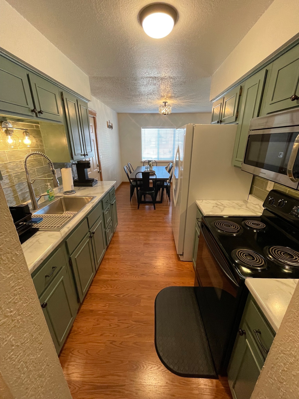 The kitchen features green cabinetry and a marble countertop, complemented by modern appliances. A long, narrow layout directs attention towards a dining area illuminated by natural light from a nearby window. The polished wooden floor enhances the space's warm atmosphere.