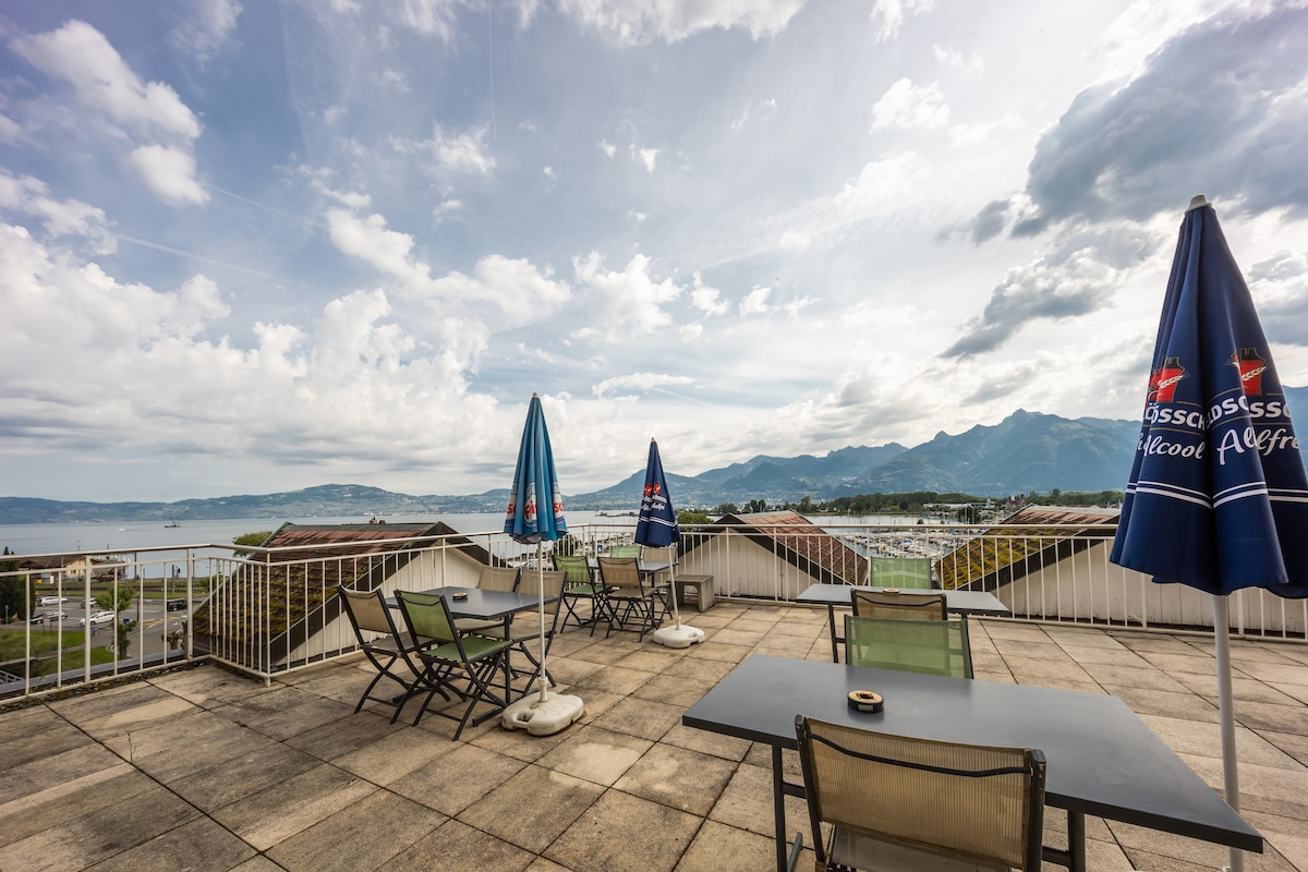 A spacious terrace is depicted, featuring several seating options, including tables and chairs. Umbrellas provide shade, while the scenic view encompasses the surrounding lake and mountains under a partly cloudy sky.