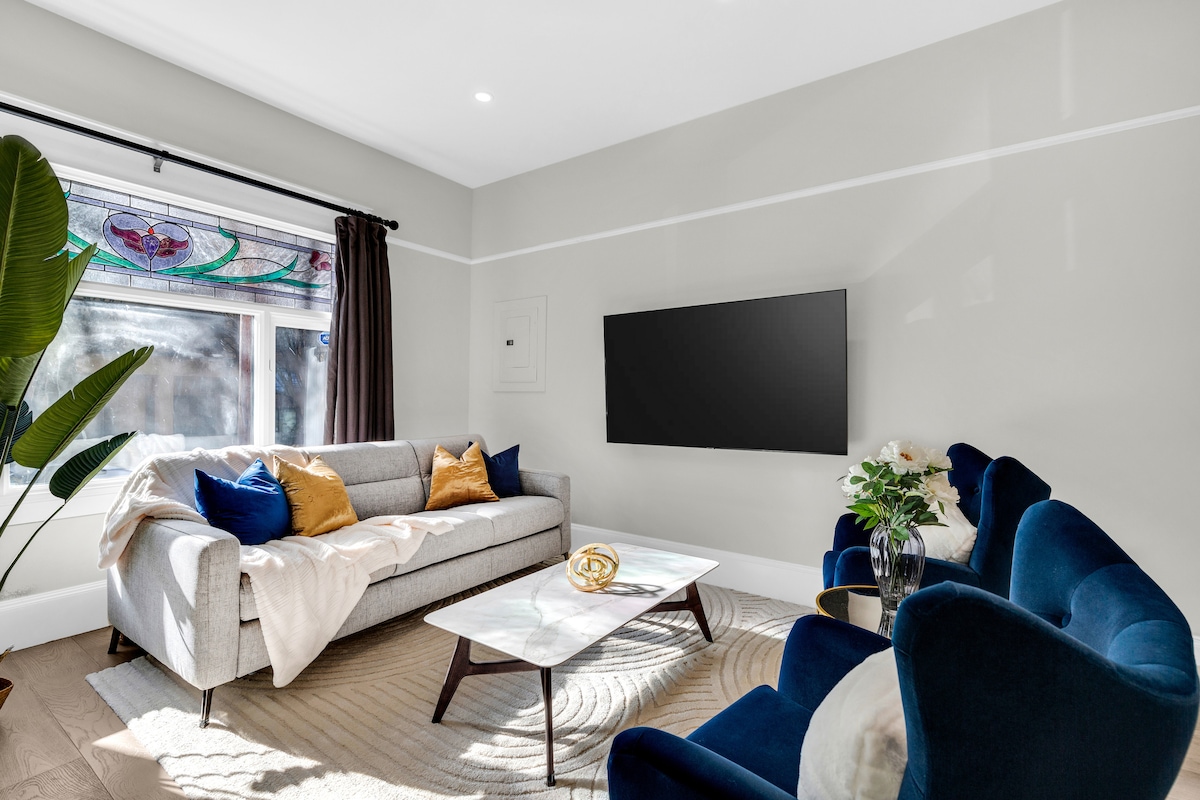 A bright living area is furnished with a contemporary gray sofa draped with a light throw and accented by colorful pillows. Two deep blue chairs complement a marble coffee table. A stained glass window adds character, while the space is softly illuminated by natural light.