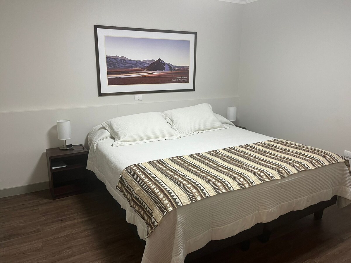 A well-appointed room features a spacious bed with a patterned throw blanket. Two bedside lamps frame the bed, and a landscape artwork is displayed above the headboard, complementing the neutral wall colors and wooden flooring.
