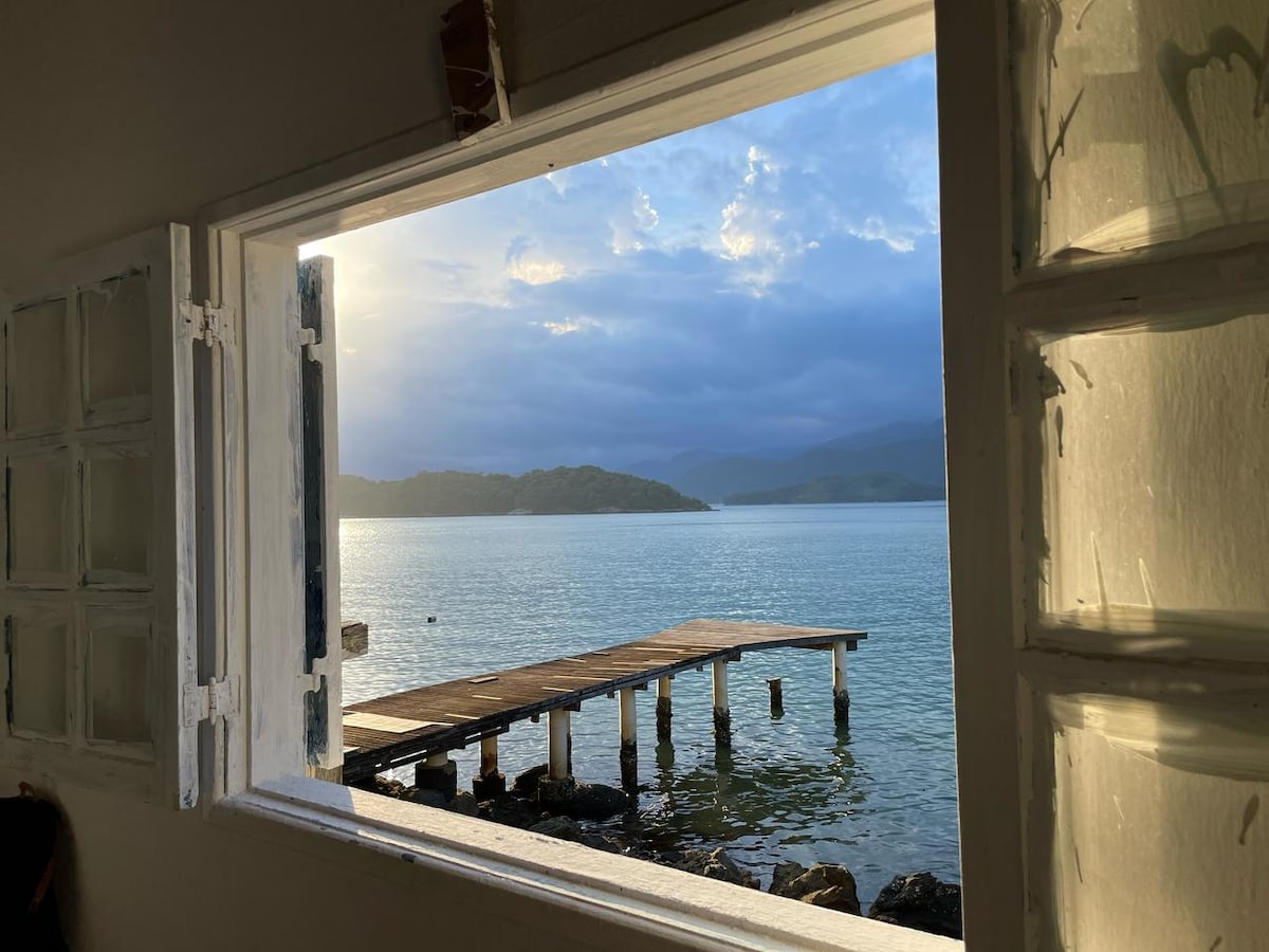 A window frames a serene view of the water, where gentle waves kiss a wooden pier jutting out into the bay. Soft clouds can be seen above, and lush green hills outline the distant shoreline. Natural light fills the interior space, enhancing the tranquil atmosphere.