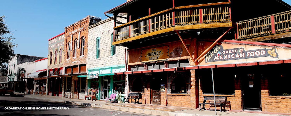 A row of historic buildings featuring a mix of stone and wood façades. A prominent balcony is visible above a restaurant with a large sign that reads 'A Great Mexican Food.' Outdoor seating areas and colorful accents contribute to the appealing storefronts.