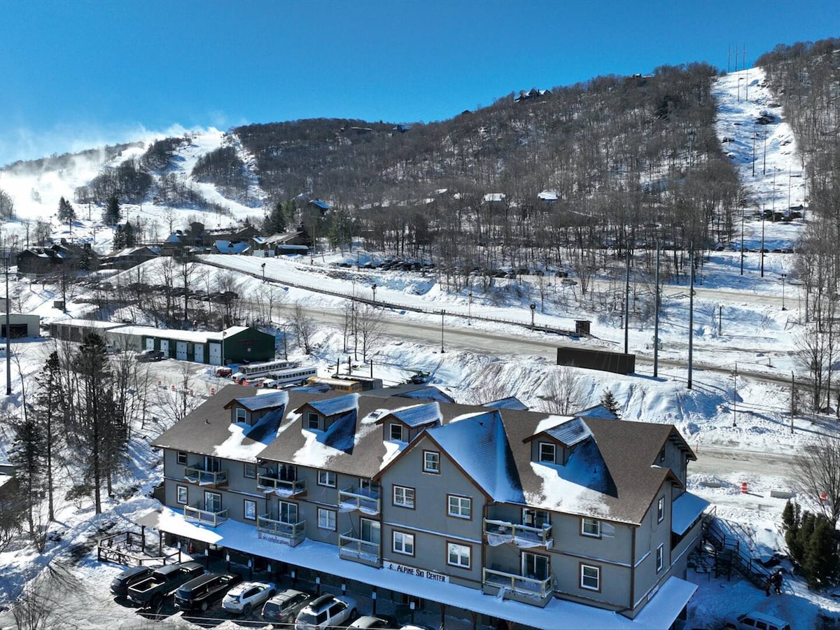 The image captures a snow-covered landscape featuring a multi-story building with a dark roof. Below, parked vehicles are visible in designated spaces. In the background, snow-covered ski slopes ascend gently, contrasted by trees and clear blue skies, creating a serene winter scene.