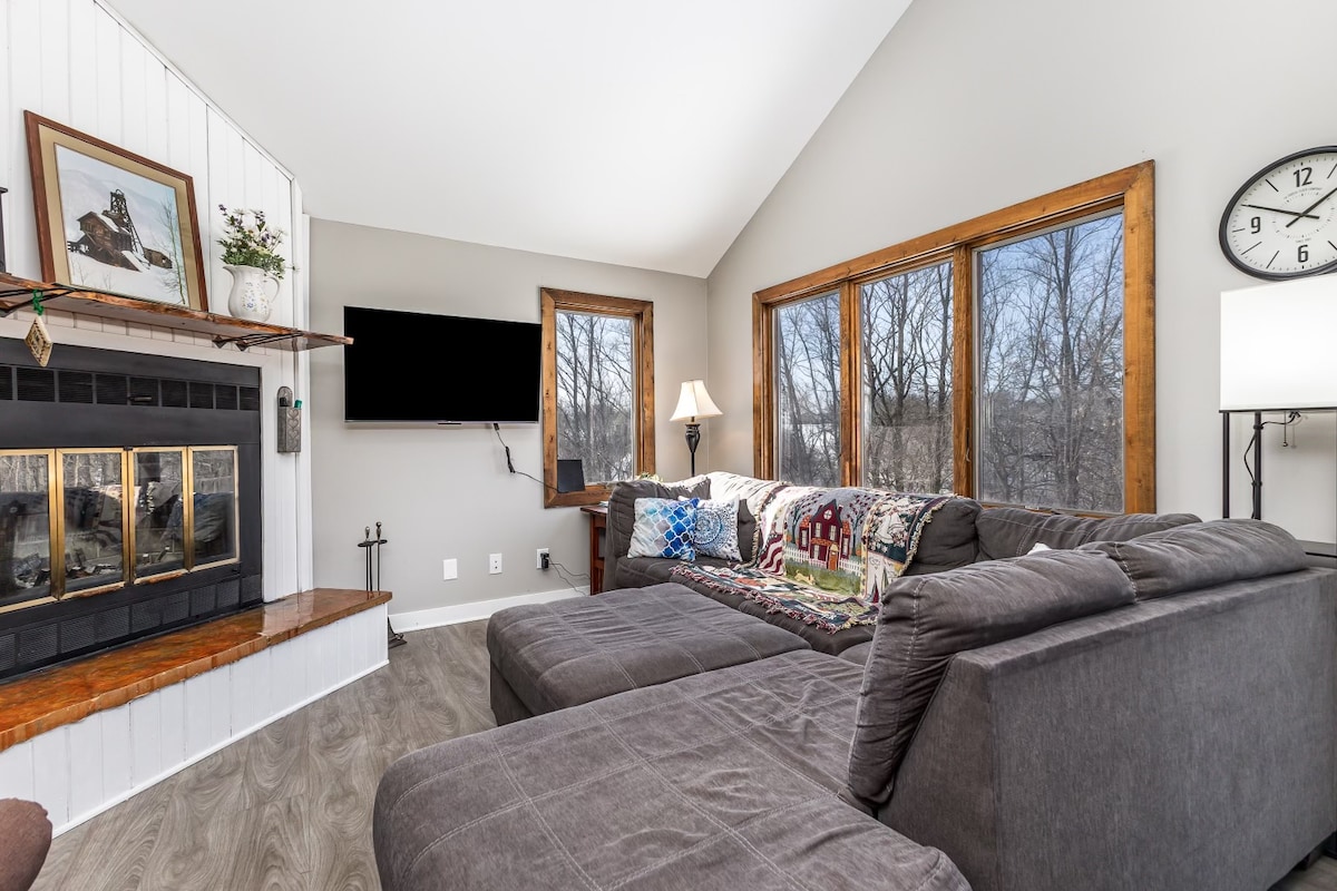 A spacious living area features a large sectional sofa adorned with decorative throw pillows. Large windows allow natural light to illuminate the space, presenting views of the wooded surroundings. A mounted television is positioned above a cozy fireplace, enhancing the room's inviting atmosphere.