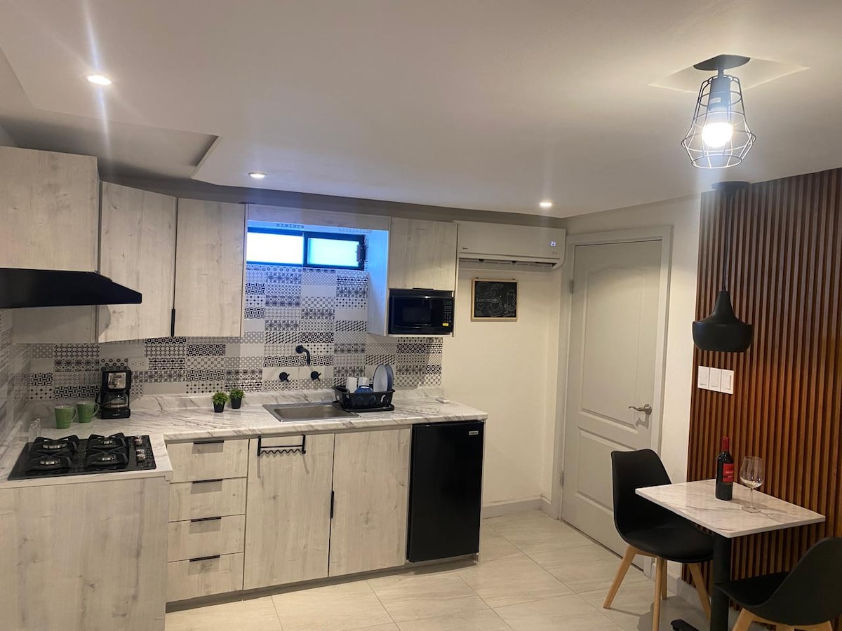A modern kitchen with light-colored cabinetry and a marble countertop features a gas stove, microwave, and sink. A small dining area with two black chairs and a round table is positioned near a window. An air conditioning unit is also visible.