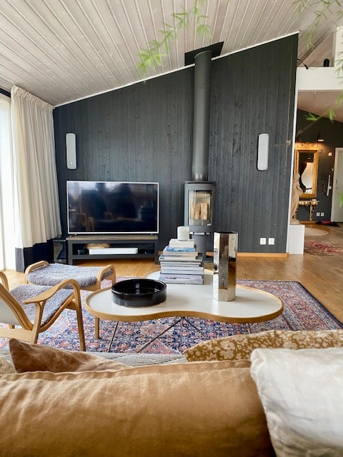 A cozy living area features a modern television atop a dark wooden cabinet. A soft sofa with neutral tones is complemented by a round coffee table that holds a stack of books and a decorative bowl. The room is warmed by a wood-burning stove against a dark wooden accent wall.