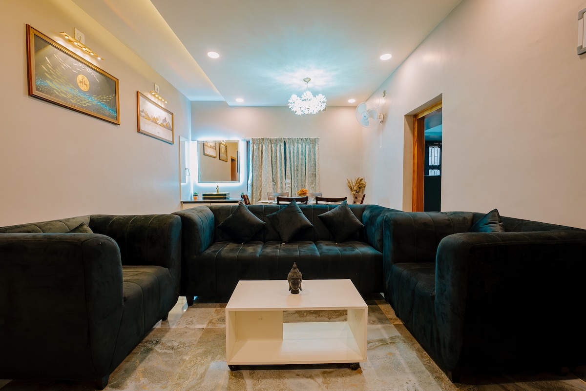 A spacious living area is depicted with a dark sofa arranged in a U-shape, complemented by a sleek white coffee table at the center. Soft lighting enhances the ambiance, while a stylish chandelier hangs overhead. A dining area is visible in the background, featuring a wooden table and elegant decor.