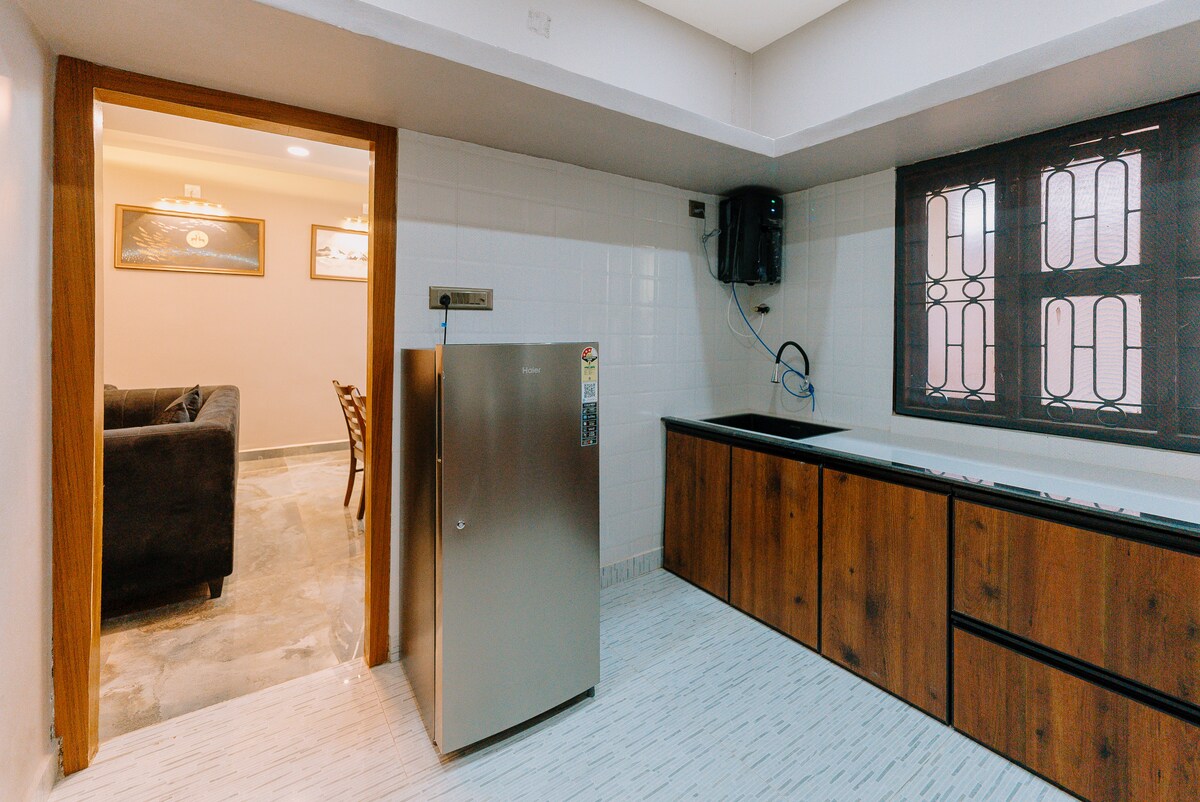 A modern kitchen is depicted with sleek wooden cabinets and a minimalist design. A stainless steel refrigerator stands prominently, complemented by a dark countertop. Natural light filters through a window with decorative ironwork, illuminating the spacious area that connects to a cozy living space.