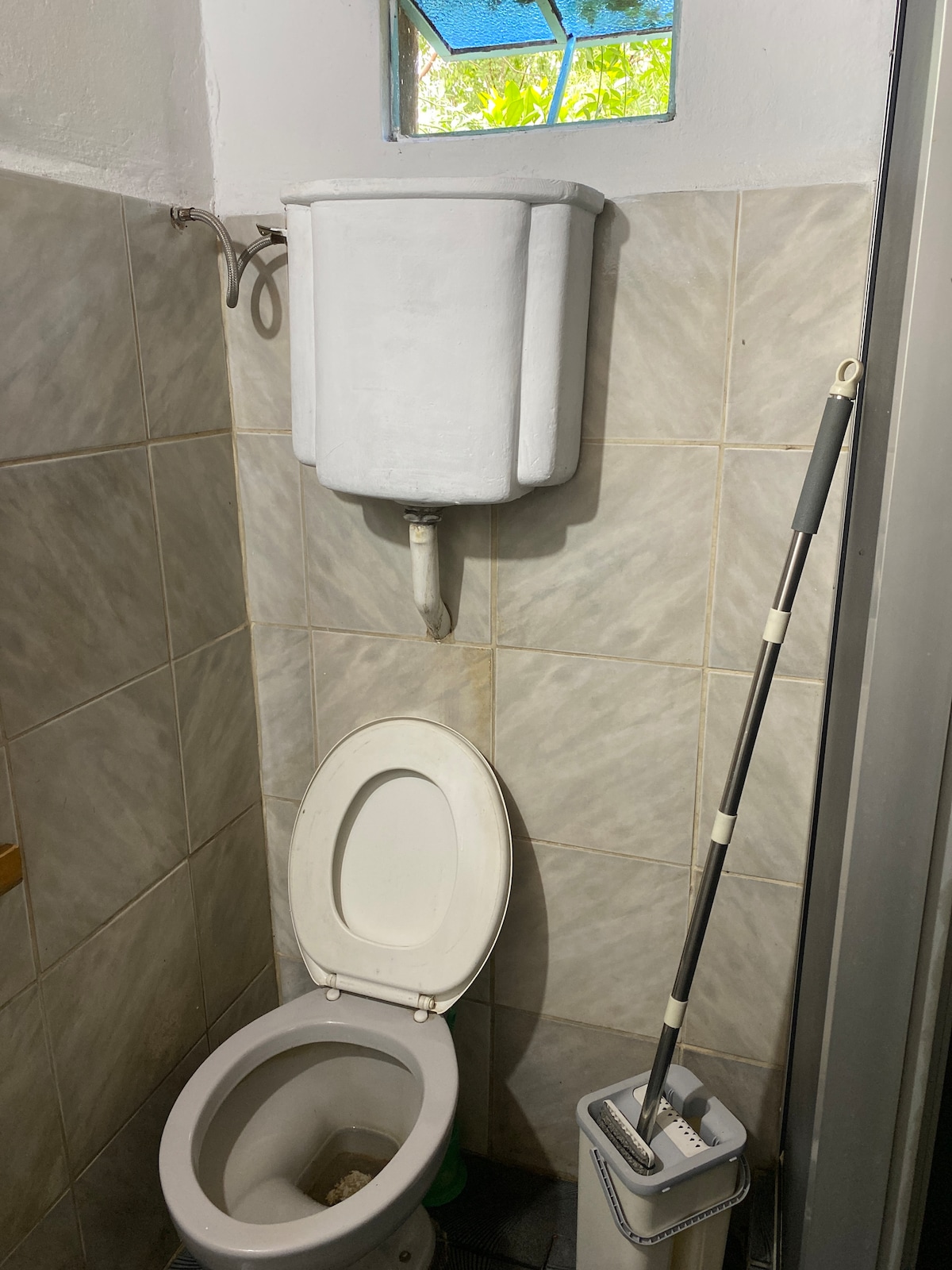 A functional bathroom space features a standard toilet positioned against tiled walls. Above the toilet, a wall-mounted water tank is visible. A cleaning brush and holder are placed nearby. Natural light enters through a small window, adding brightness to the area.