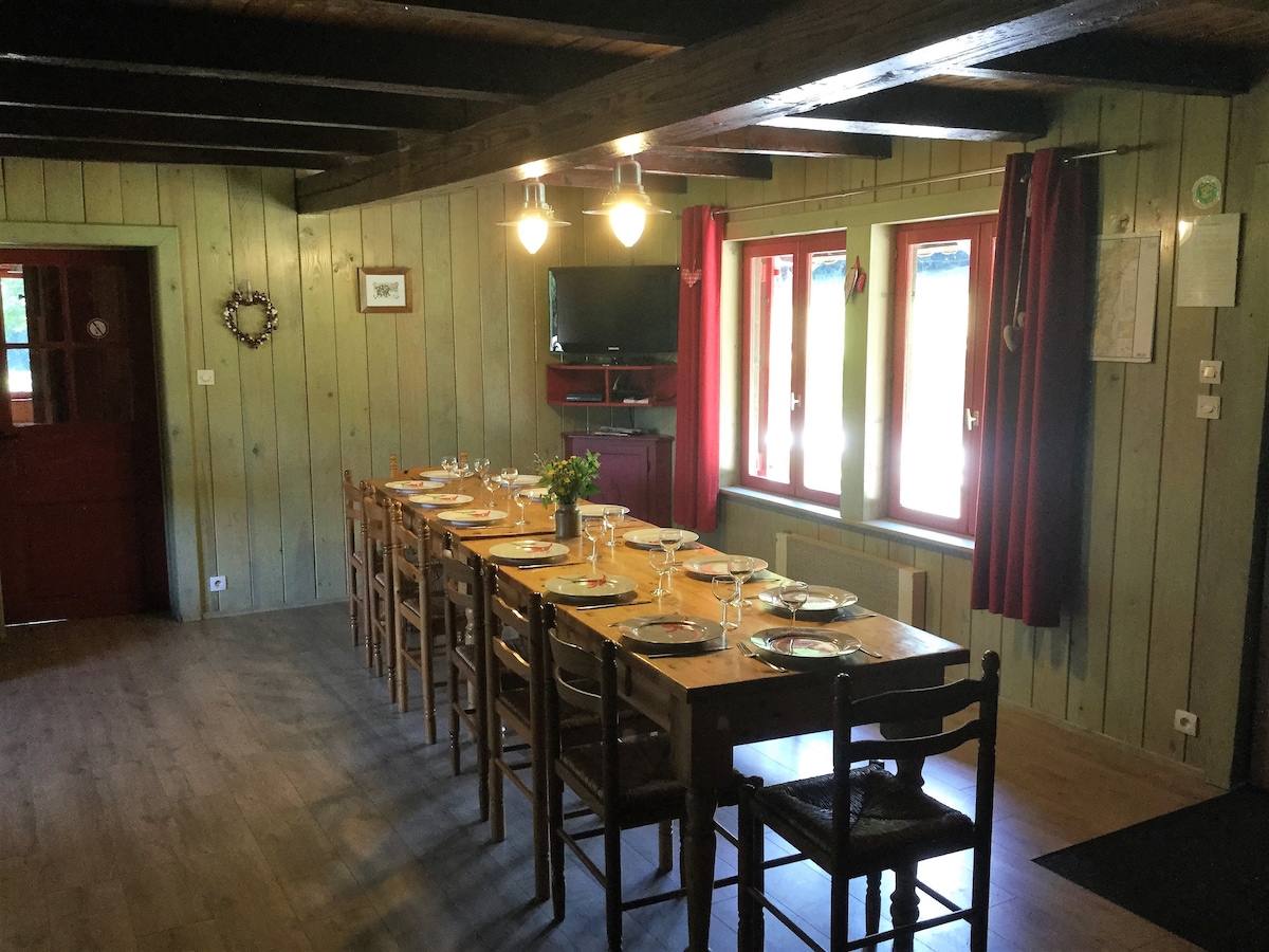 A spacious dining area features a long wooden table set for a meal, surrounded by wooden chairs. Large windows allow natural light to illuminate the room, complemented by wooden beams on the ceiling. A television is mounted on the wall, contributing to the cozy atmosphere.