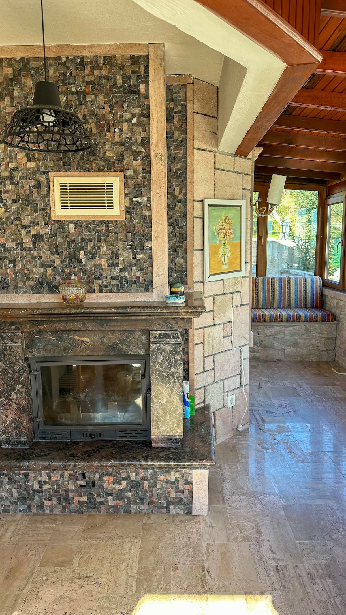 A stone fireplace is framed by decorative stonework, enhancing a cozy corner in a sunlit enclosed veranda. Natural light is allowed in through multiple windows, while a colorful striped seating area invites relaxation. Wooden beams highlight the ceiling, adding warmth to the space.