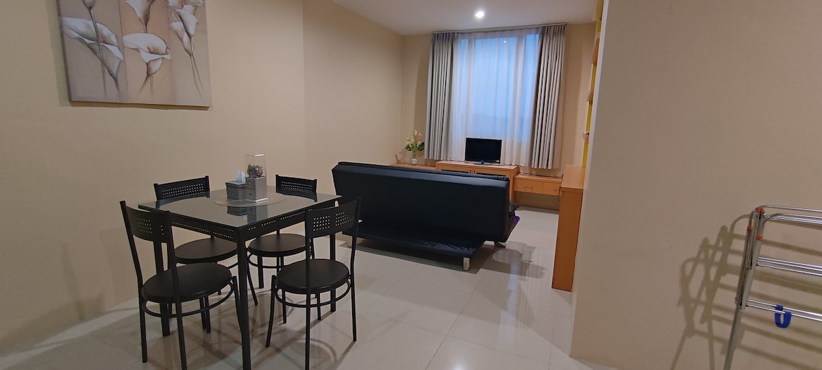 A dining area is depicted with a clear glass table and four black chairs. Adjacent, a dark sofa faces a wooden media console with a television. Natural light filters in through a light, sheer curtain covering a window, enhancing the neutral tones of the walls.