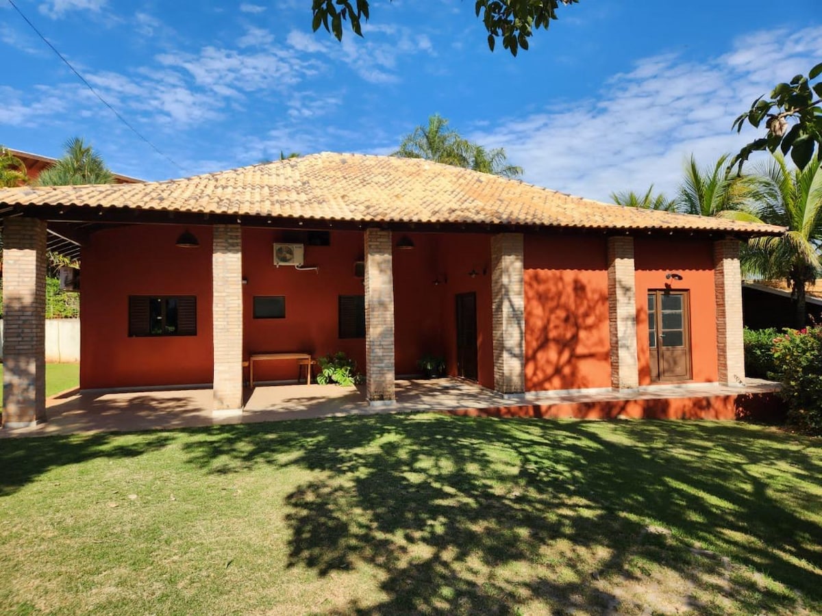 A broad façade of a house is highlighted, featuring a terracotta exterior and a tiled roof. The structure is supported by brick columns. Windows with dark shutters are positioned symmetrically, and greenery surrounds the house, creating a relaxed outdoor setting.