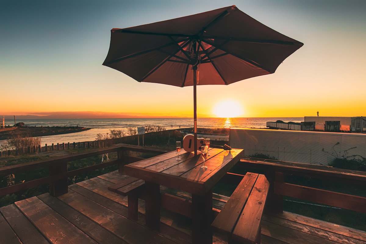 A wooden patio table with an umbrella is positioned to capture a breathtaking sunset view over the beach. The scene features a calm ocean reflecting shades of orange and yellow, with silhouettes of nearby structures enhancing the tranquil atmosphere.