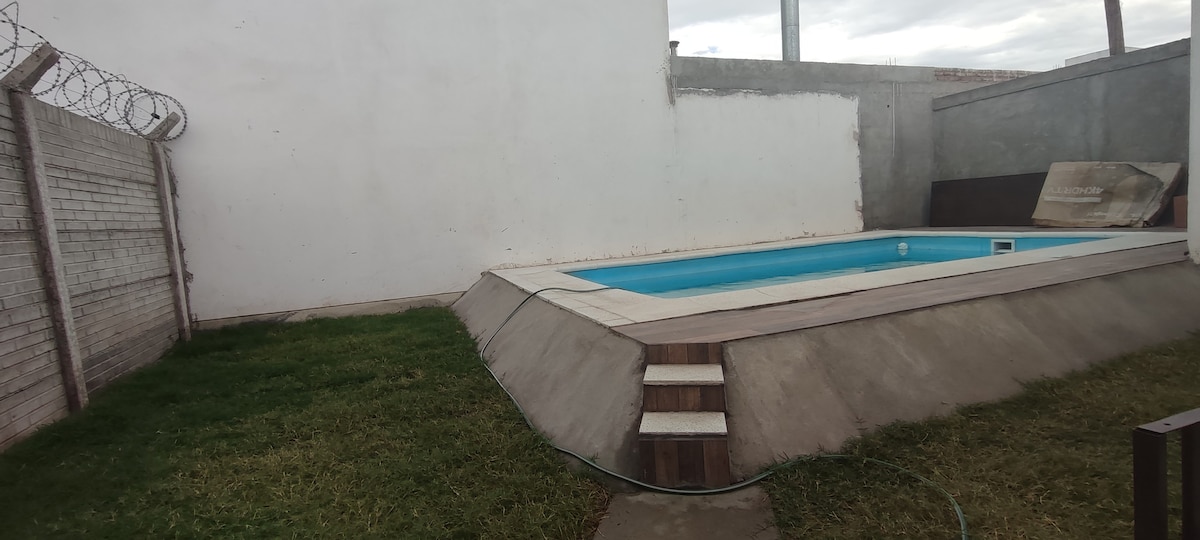 A rectangular swimming pool is surrounded by a smooth concrete deck, featuring steps on one side leading into the water. An expanse of grass borders the pool, with a bare white wall in the background. A fence with barbed wire is visible along the side.