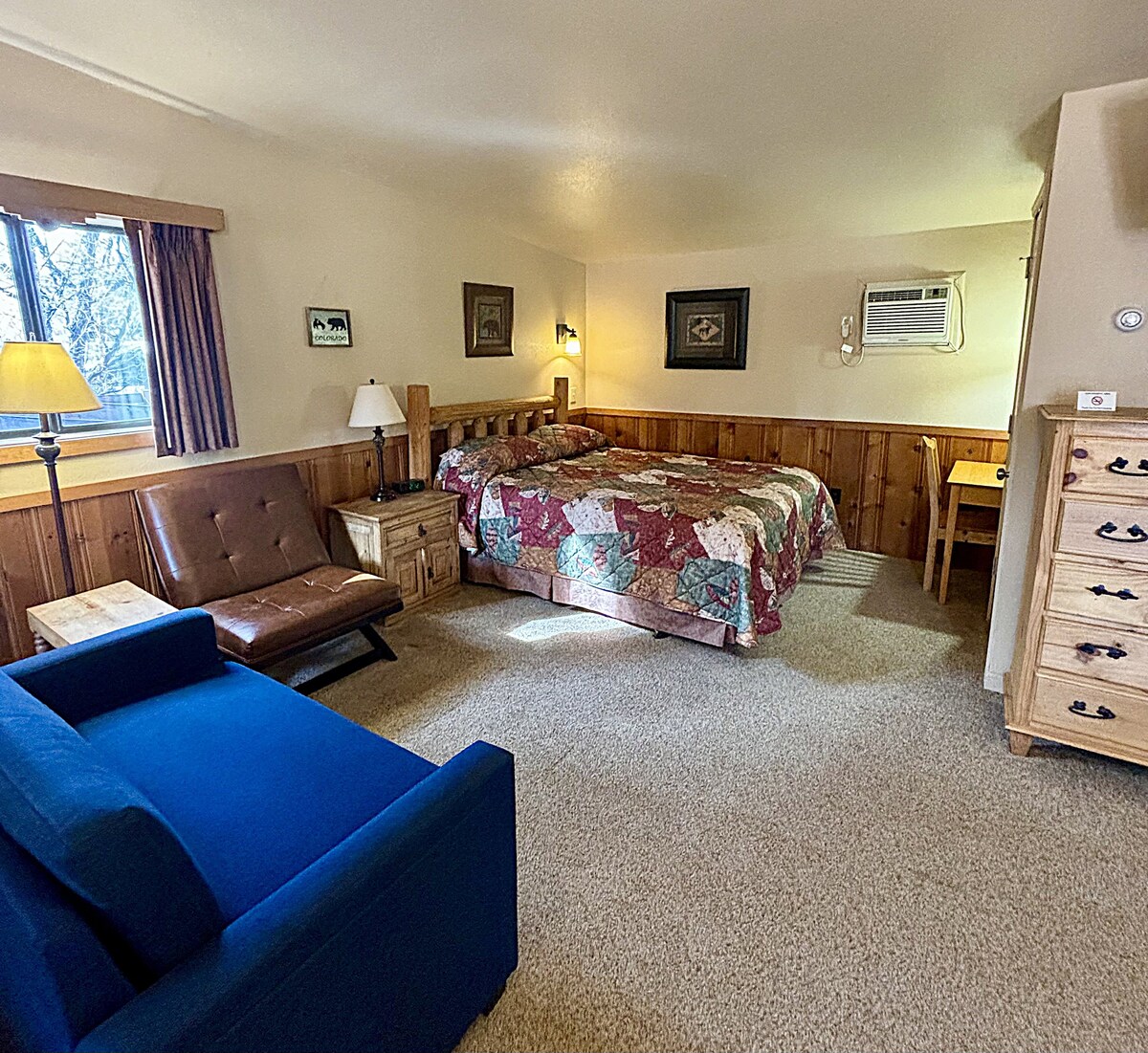 A cozy guest room features a spacious bed covered with a patterned quilt, complemented by wooden accents. A blue sofa and a small dining table are present, while natural light enters through a window with light curtains. An air conditioning unit is mounted on the wall.