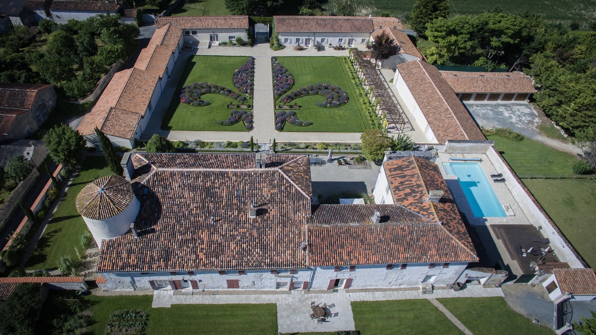 An aerial view captures the rustic charm of a sprawling estate surrounded by meticulously manicured gardens featuring a decorative floral design. A swimming pool is visible to the right, while the historic buildings showcase traditional red-tiled roofs, blending harmoniously with the lush green landscape.