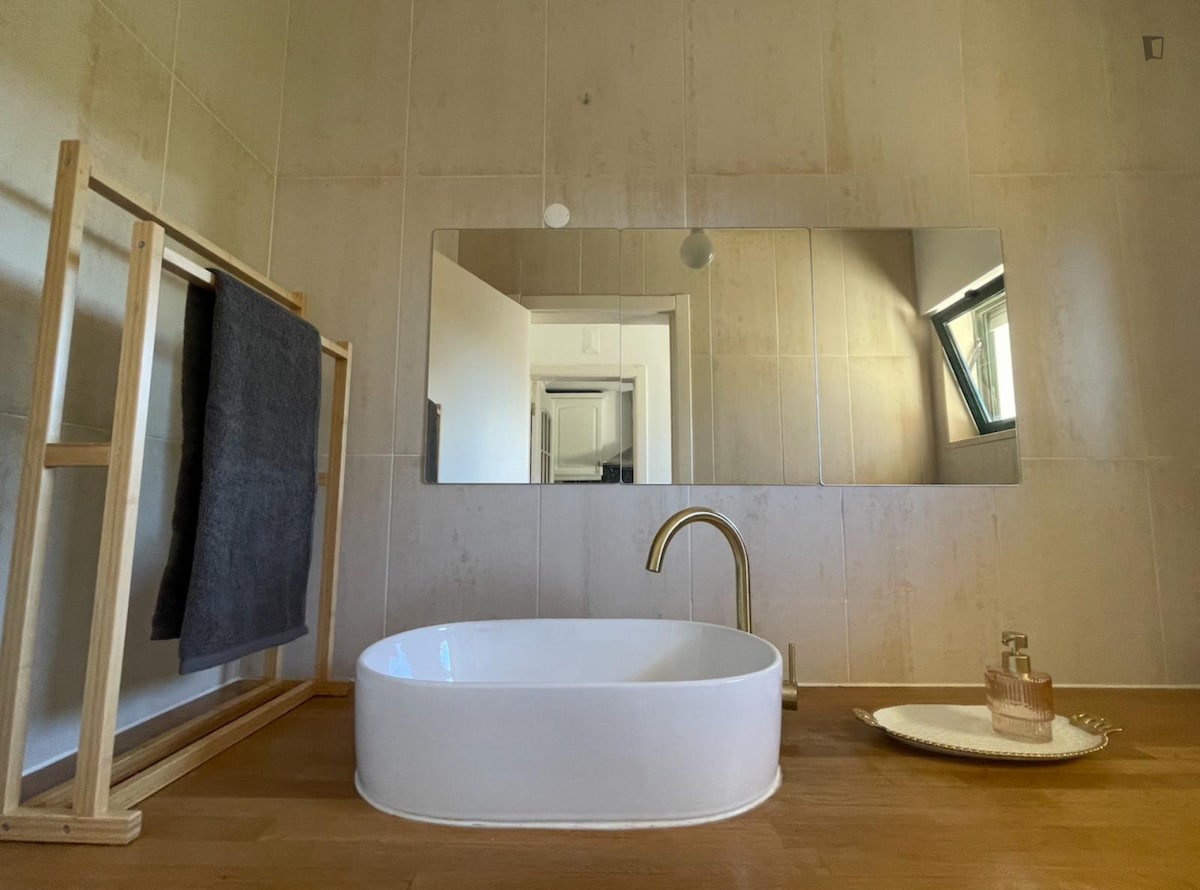 A modern bathroom is presented with a freestanding oval sink positioned in front of a large, frameless mirror. A wooden rack holds a gray towel, while a small tray displays a bottle and a decorative item on the countertop.