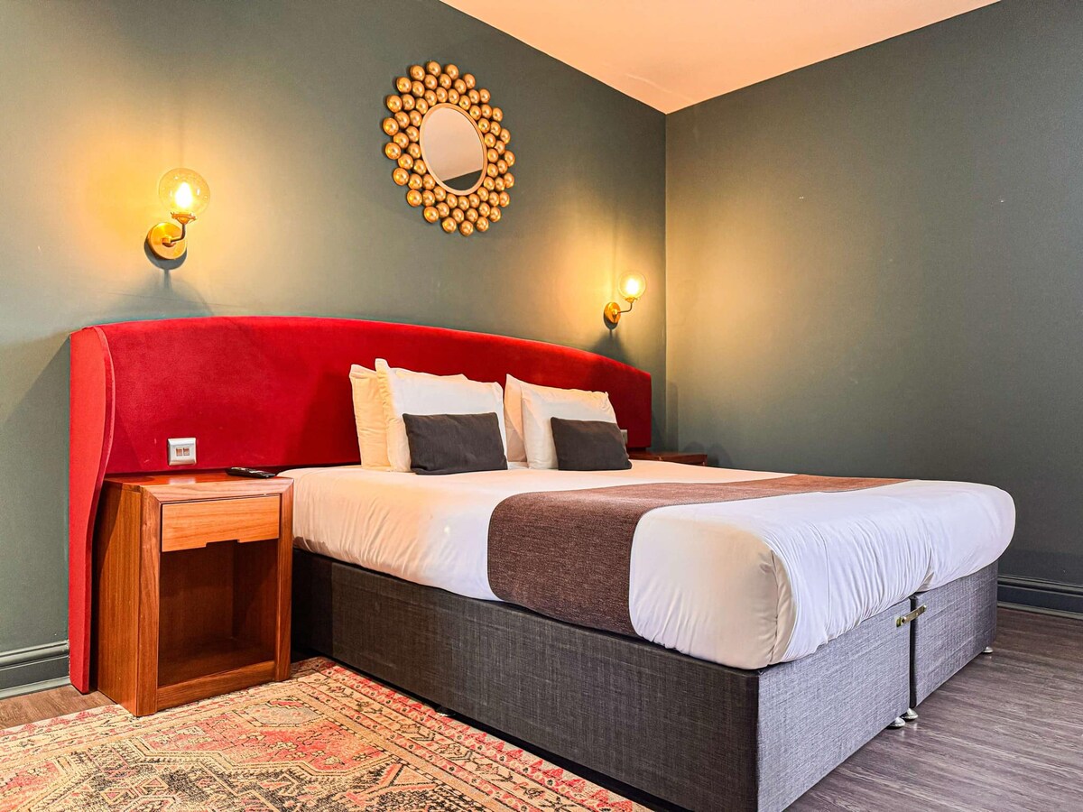 The image shows a spacious bed with a dark headboard and a vibrant red accent wall. Two bedside wall sconces provide soft lighting. A small wooden nightstand is positioned beside the bed, enhancing the room's design. A decorative mirror is mounted above the headboard, reflecting the ambiance.