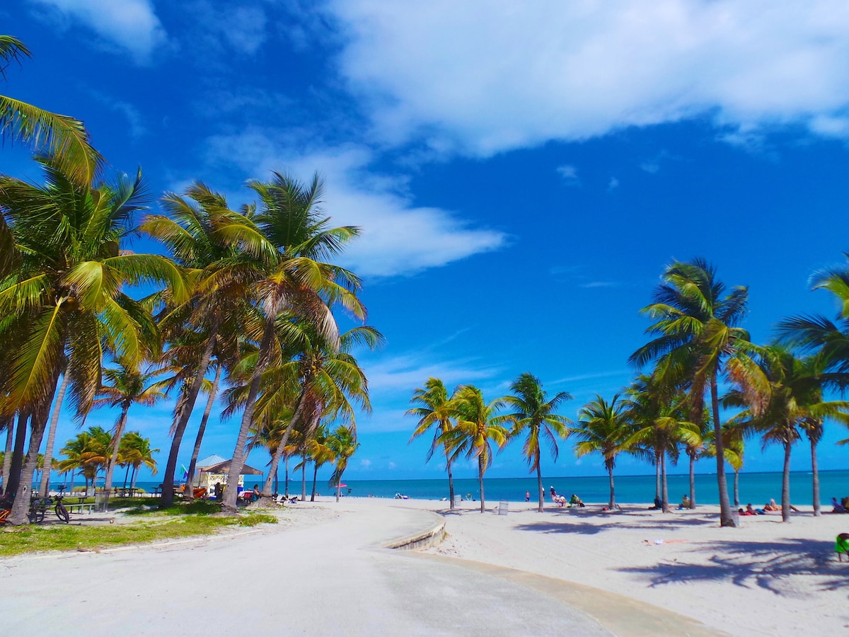 A sandy beach is lined with tall palm trees, swaying gently in the breeze. A clear blue sky stretches overhead, while the tranquil ocean is visible in the background. Sun loungers and small umbrellas are scattered along the shore, inviting relaxation.