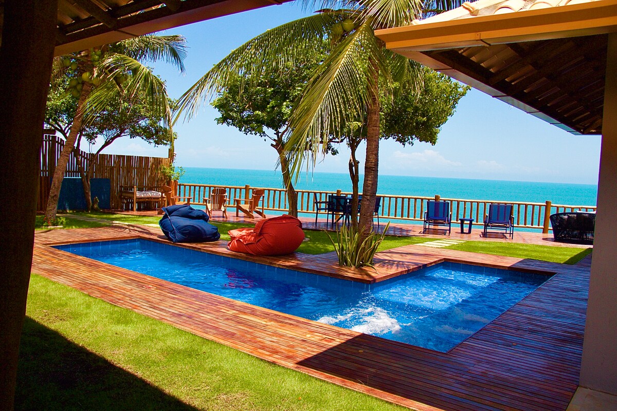 A swimming pool is framed by wooden decking and a lush green lawn, with two colorful bean bag chairs positioned nearby. Palm trees sway in the gentle breeze, and an expansive view of the turquoise ocean is visible beyond the railing.
