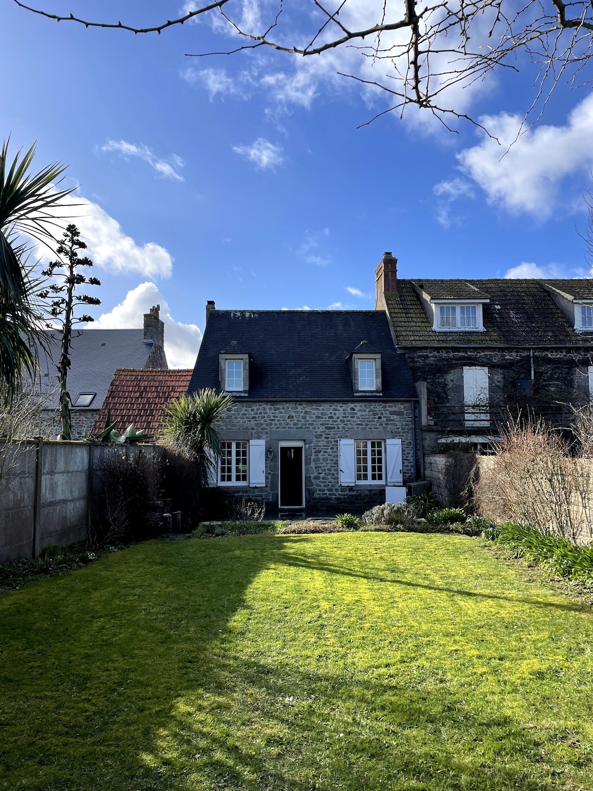 A charming house with a stone facade is framed by a neatly maintained garden. The front door is centered under an overhang, while large windows allow natural light to enter. Lush green grass stretches across the yard, and structures are visible on either side.