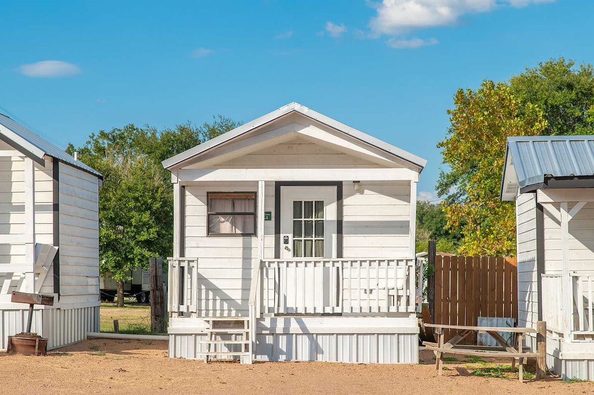 A charming log cabin is depicted, featuring a covered porch with a simple bench. The cabin is painted in a light color, with a door and window visible on the front. Surrounding trees and clear blue skies create a serene outdoor setting.