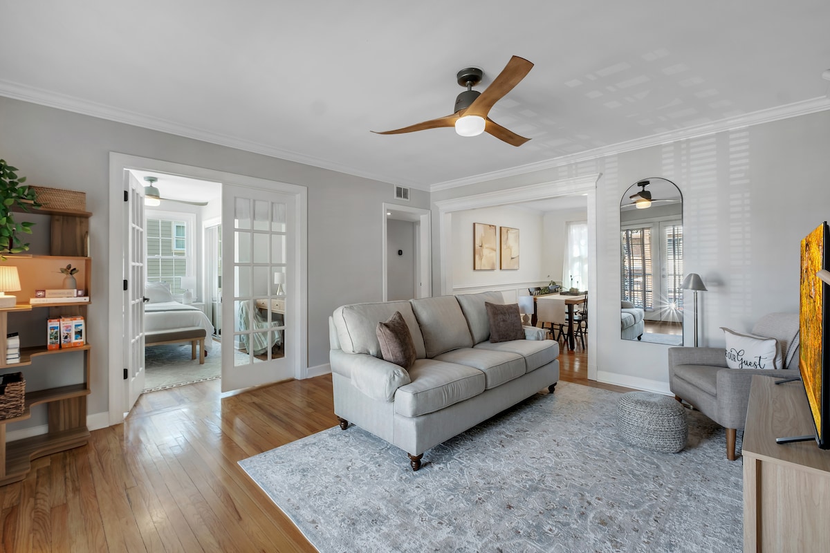A spacious living area is centered around a light gray sofa, complemented by a sleek ceiling fan. Natural light streams in through clear windows, highlighting the warm hardwood floors. A large area rug defines the seating space, while a mirrored wall adds depth to the room.