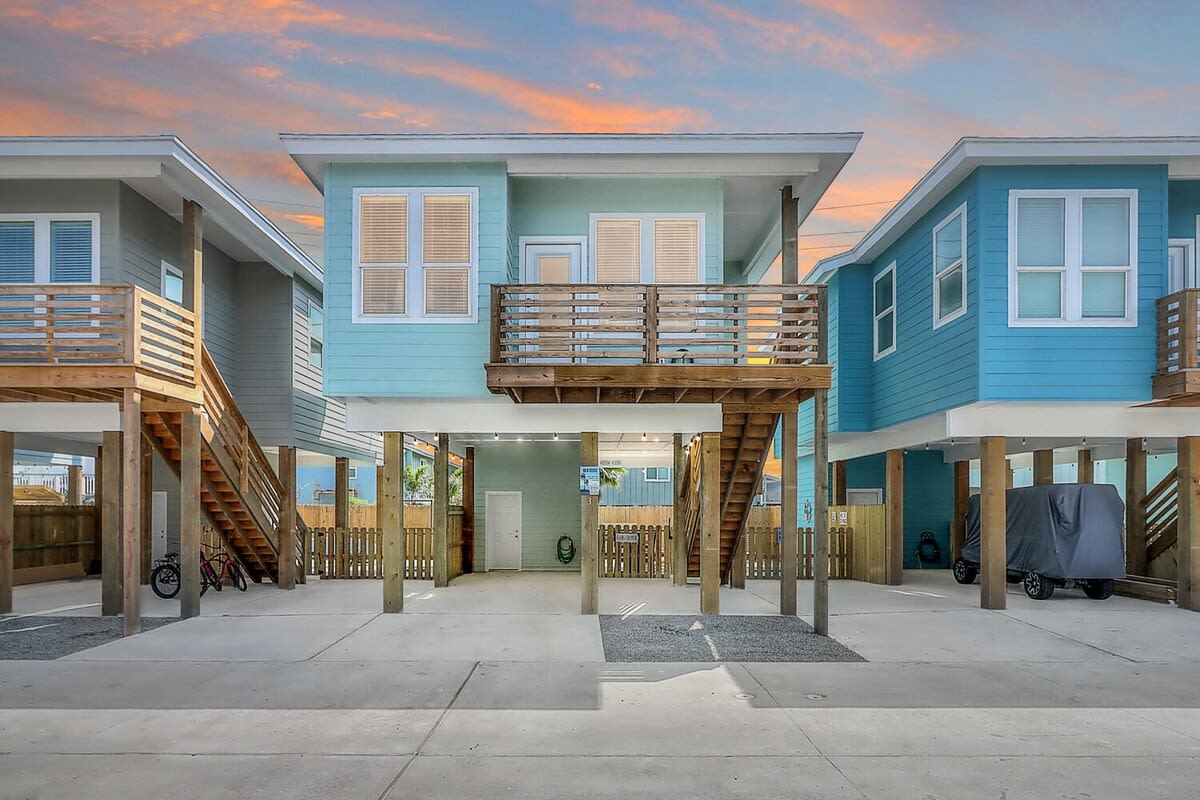 A beach house design features a two-story facade with light blue siding and multiple balconies. The image captures a symmetrical layout with wooden railings and steps leading to the entrances. The outdoor area is paved and lined with wooden fencing, offering a welcoming atmosphere.