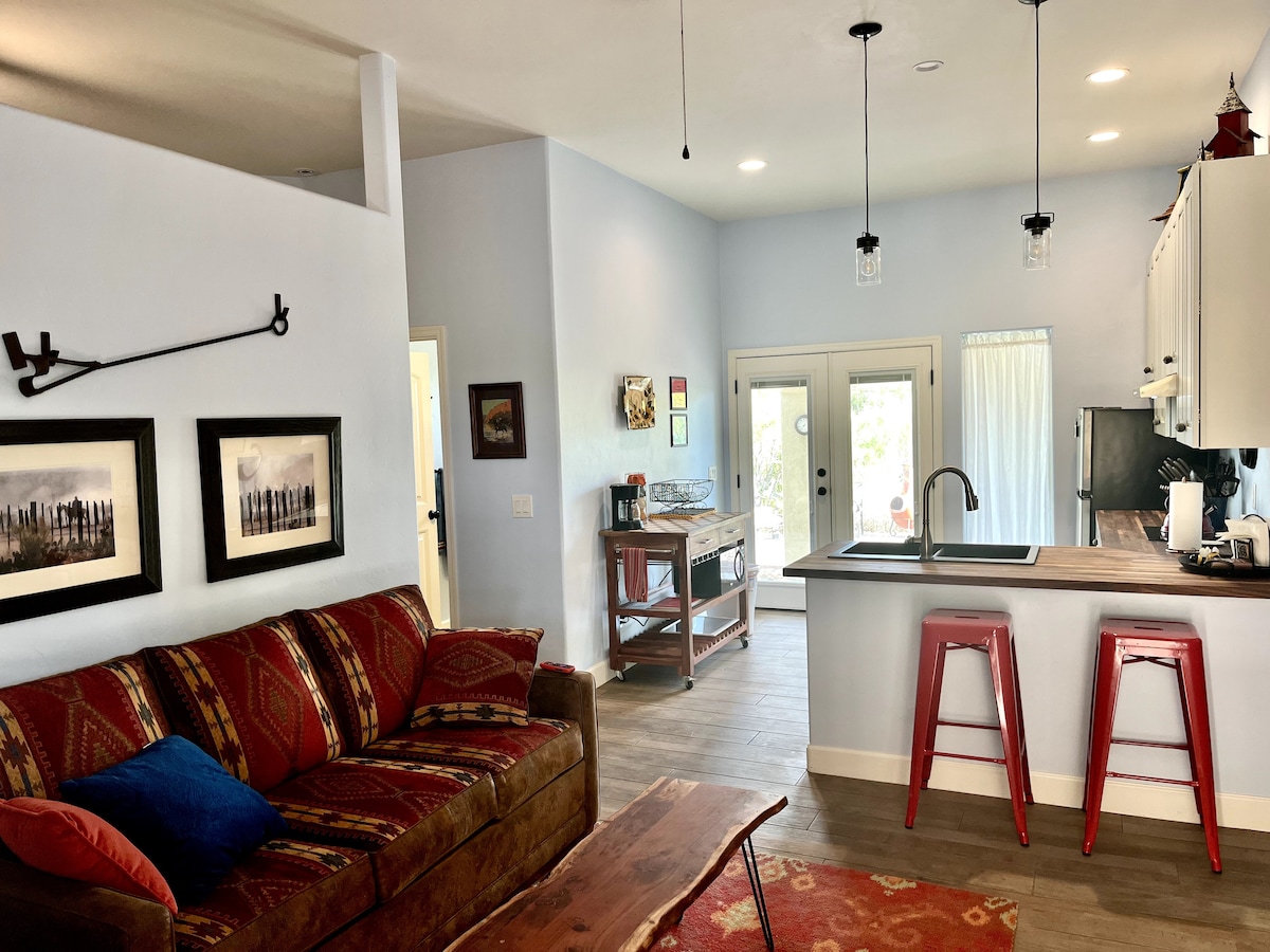An open living area is depicted, featuring a comfortable sofa arranged with decorative pillows. The kitchen area includes a countertop with stools and hanging light fixtures. Sliding glass doors allow natural light and views of the outdoors, enhancing the inviting ambiance of the space.