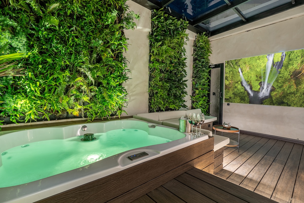 A private terrace features a modern jacuzzi surrounded by lush vertical greenery. A small table with glasses and a bottle stands nearby, creating a serene atmosphere. A large wall screen displays an image of a peaceful nature scene, enhancing the relaxing outdoor space.