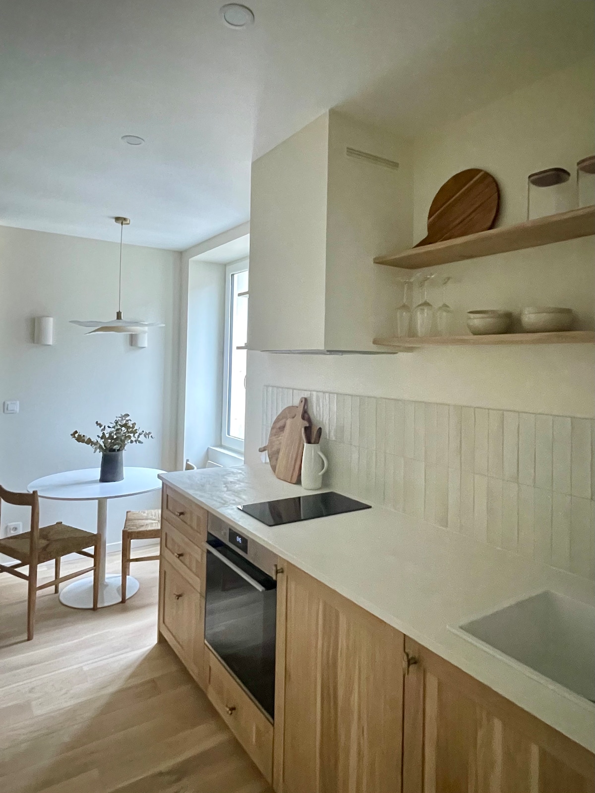 The kitchen area features light-colored cabinetry and a white countertop with an integrated sink. A round dining table is set for two, accompanied by a wooden chair. Open shelving displays kitchenware, while a pendant light hangs above, illuminating the space.
