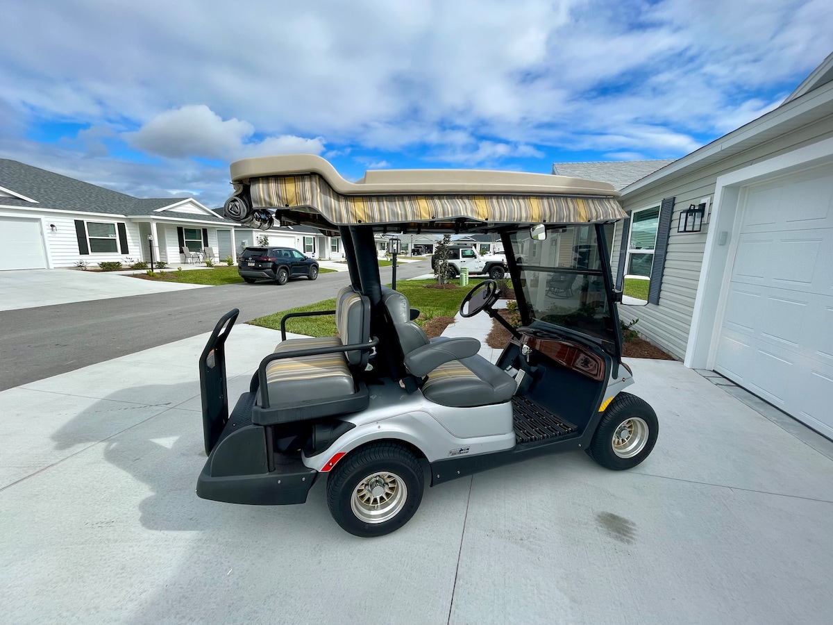A four-seater golf cart is positioned on a concrete driveway, featuring a beige and gray canopy overhead. The cart has comfortable seating and is equipped with a steering wheel and dashboard controls, set against a backdrop of nearby homes and a clear blue sky.