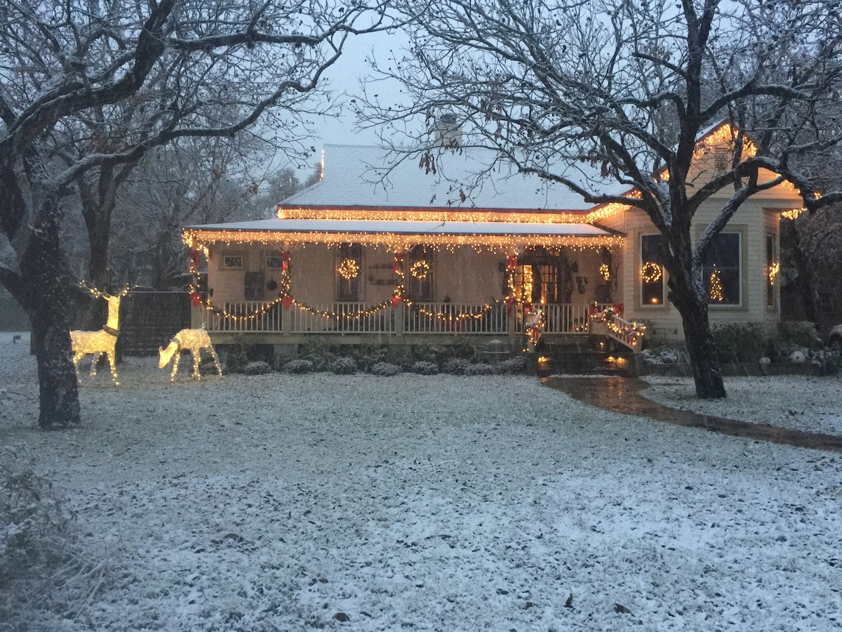 A charming house decorated with festive lights is framed by bare trees in a snow-covered yard. Two illuminated reindeer figures are positioned in front, while wreaths adorn the windows. A pathway leads to the inviting entrance, creating a cozy holiday scene.