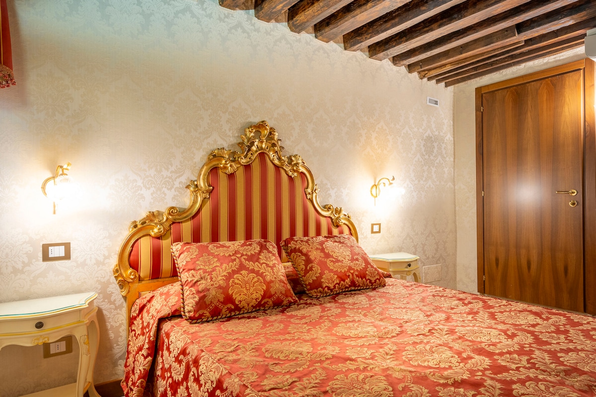 A sleeping area is highlighted by a large ornate headboard with gold detailing and red striped fabric. The bed is dressed with matching red and gold bedding, complementing the delicate wall patterns and wooden ceiling beams. Soft lighting is provided by sconces on either side.