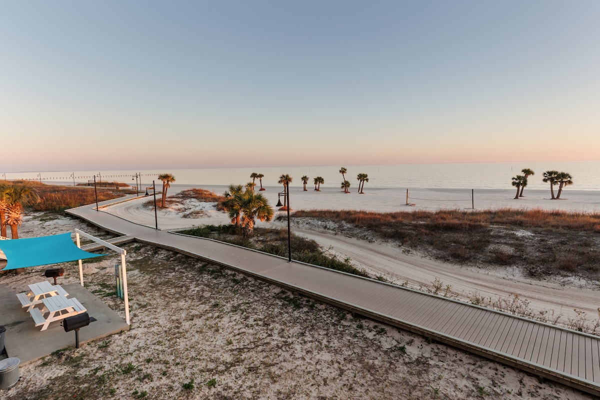 A serene beachfront view reveals a sandy path lined with palm trees, leading to the water's edge. The beach is tranquil, with gentle waves lapping at the shore under a pastel sky as the day begins to transition into evening.