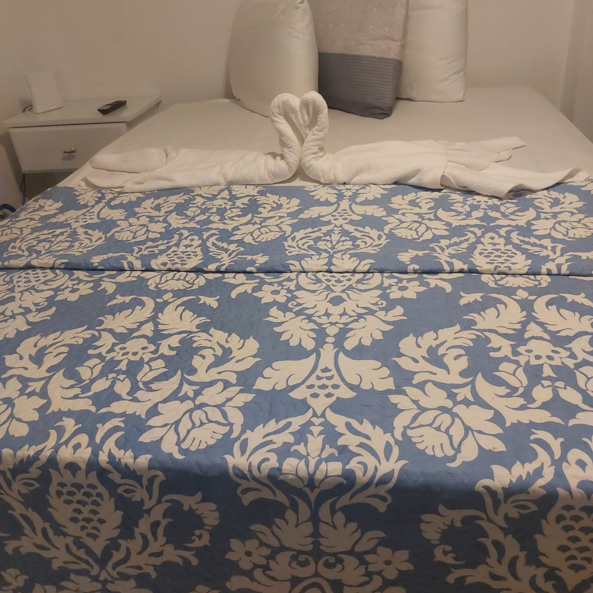 A spacious bed is dressed in a light blue and white patterned duvet, with a pair of decorative pillows placed at the head. White towels are elegantly folded into a swan shape atop the bed. A bedside table with a small lamp is visible to the side.