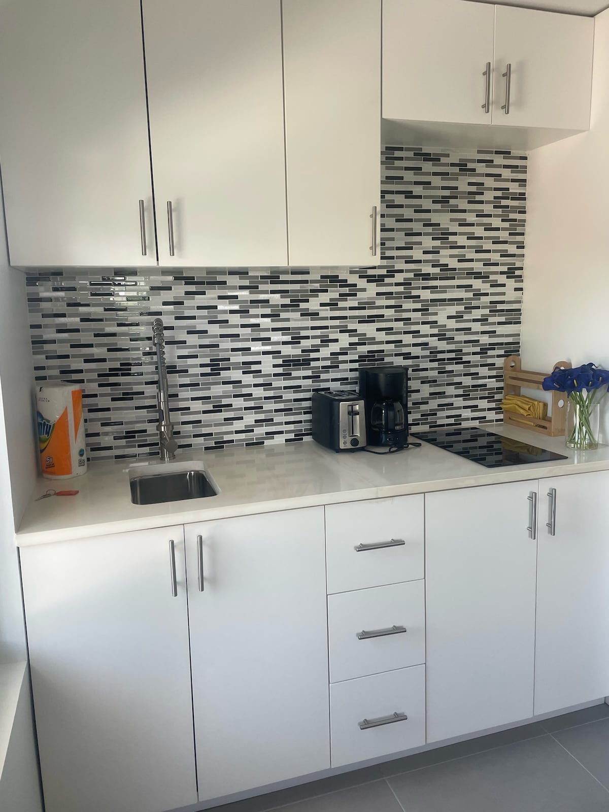 A sleek kitchenette features modern cabinetry in white, with a mosaic backsplash of black and gray tiles. Essential appliances include a coffee maker and a compact stove, complemented by a small sink. Fresh flowers add a touch of colour to the counter space.