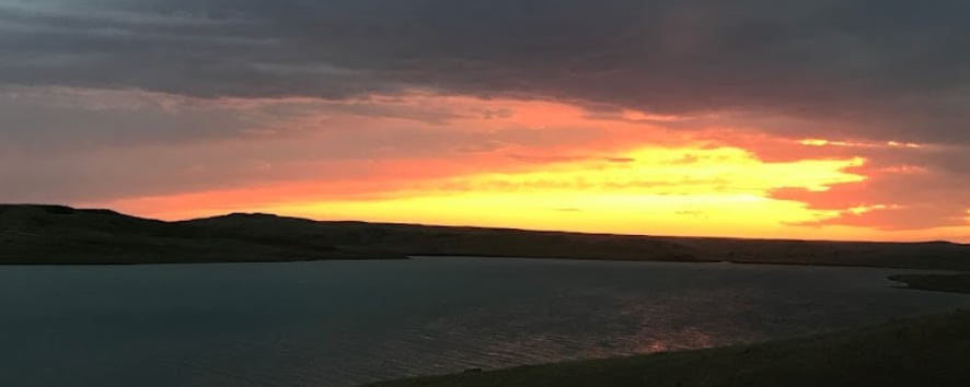 A serene sunset over Lake Oahe is captured, showcasing a gradient of warm colors in the sky. The calm waters reflect the hues of orange and pink, while the surrounding landscape features gentle hills under a softly illuminated sky.