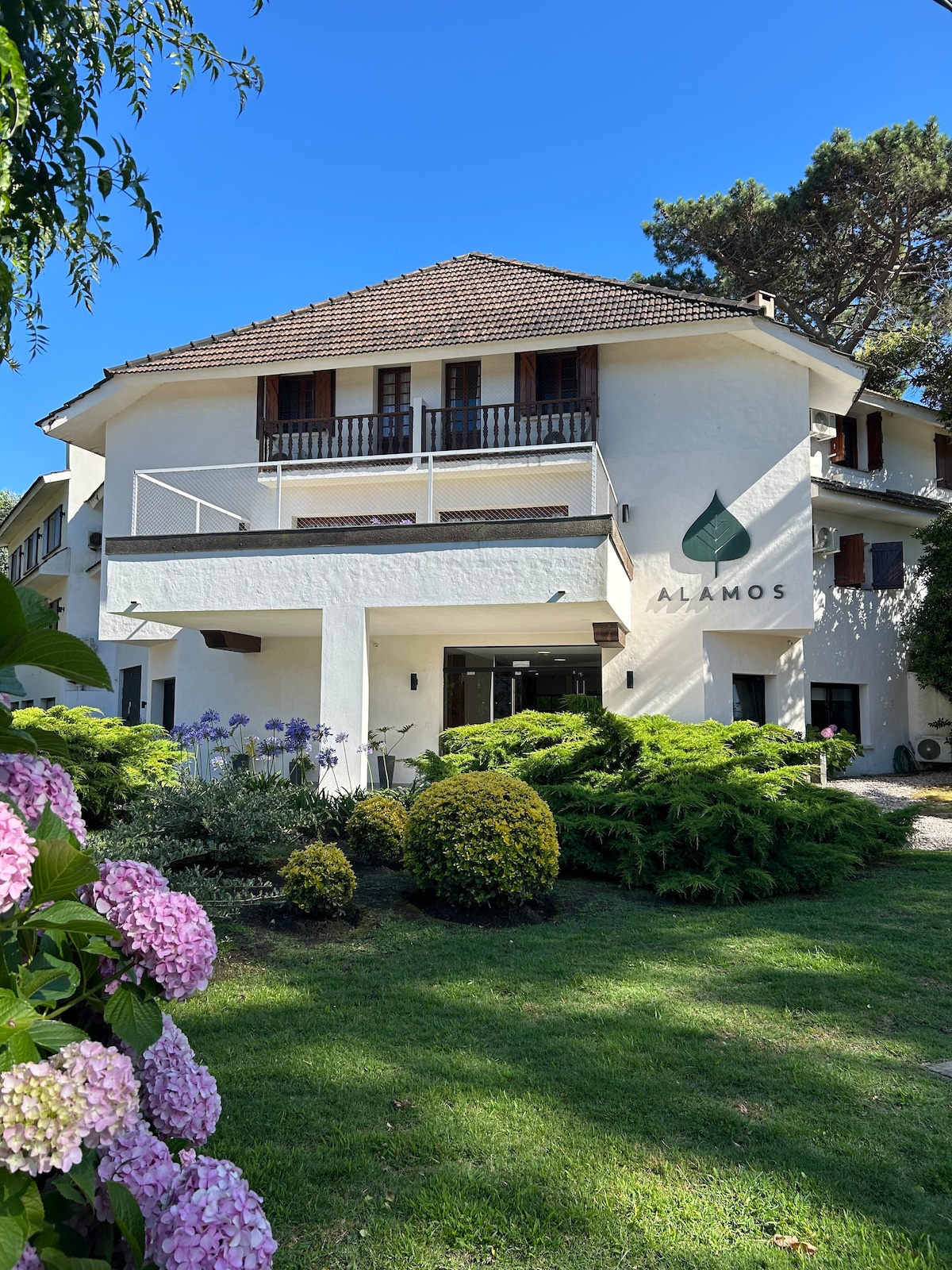 The property exterior features a multi-story building with a welcoming entrance. Well-maintained gardens with vibrant flowers and lush greenery surround the structure. A prominent logo on the facade is visible, enhancing the overall aesthetic of the location.