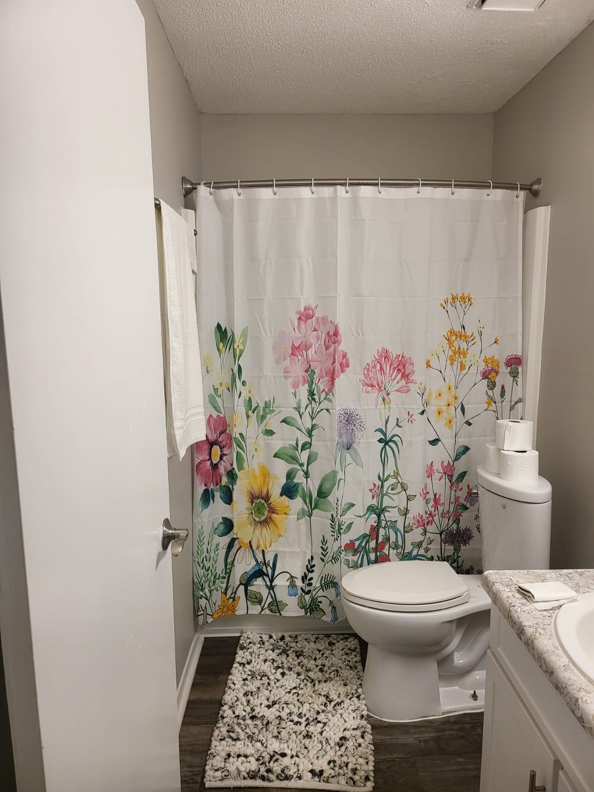 A bright bathroom is showcased with a colorful floral shower curtain that features various flowers. A white toilet is positioned next to a light-colored vanity with a countertop. A small rug lies on the floor, complementing the overall decor.
