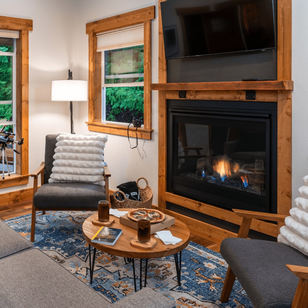 The cozy living area features a warm gas fireplace framed by wood accents. Two armchairs are positioned beside a circular coffee table, which holds snacks and beverages. A wall-mounted TV sits above the fireplace, and natural light filters through the nearby windows.