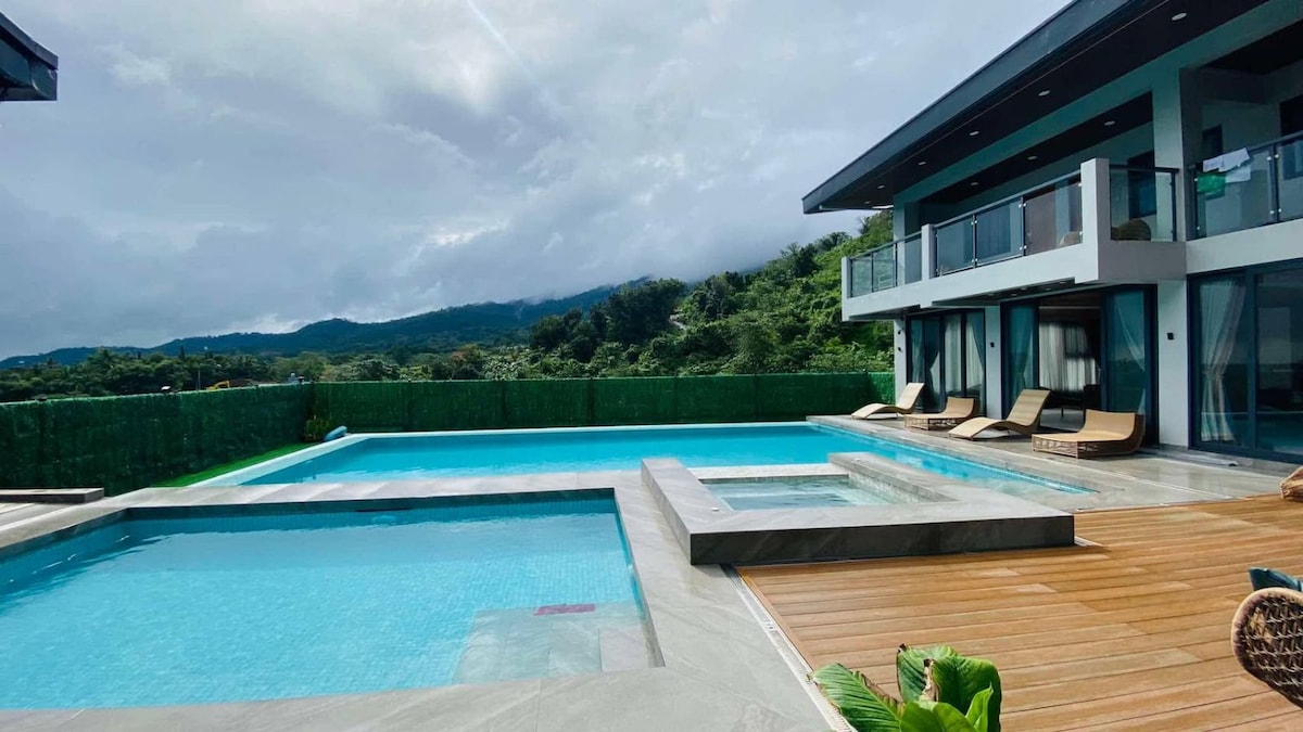 An inviting hot spring pool is showcased, featuring a large main pool and a smaller hot tub area. Sun loungers are arranged nearby on a wooden deck, with lush greenery and distant mountainous views framing the serene environment.