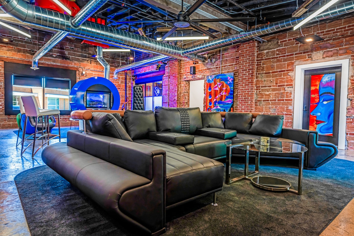 A modern lounge area features a large black sectional sofa positioned around a glass coffee table. Exposed brick walls add character, while colorful artwork and ambient lighting create a dynamic atmosphere. A bar-height table and chairs are visible in the background.