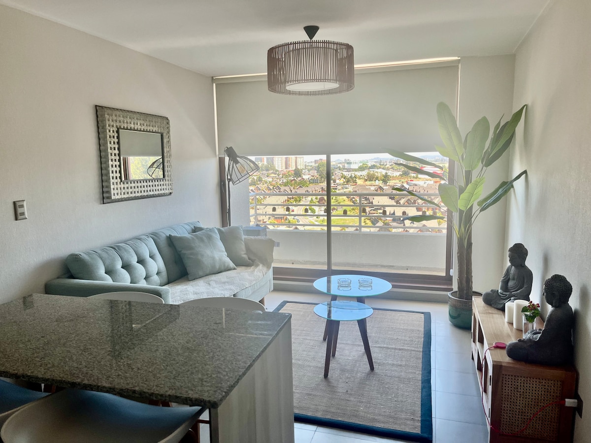 A bright living area features a comfortable light-colored sofa and a coffee table, surrounded by decorative plants. Large floor-to-ceiling windows provide a view of the outside, while a modern light fixture hangs from the ceiling. A dining area with a granite table is visible.