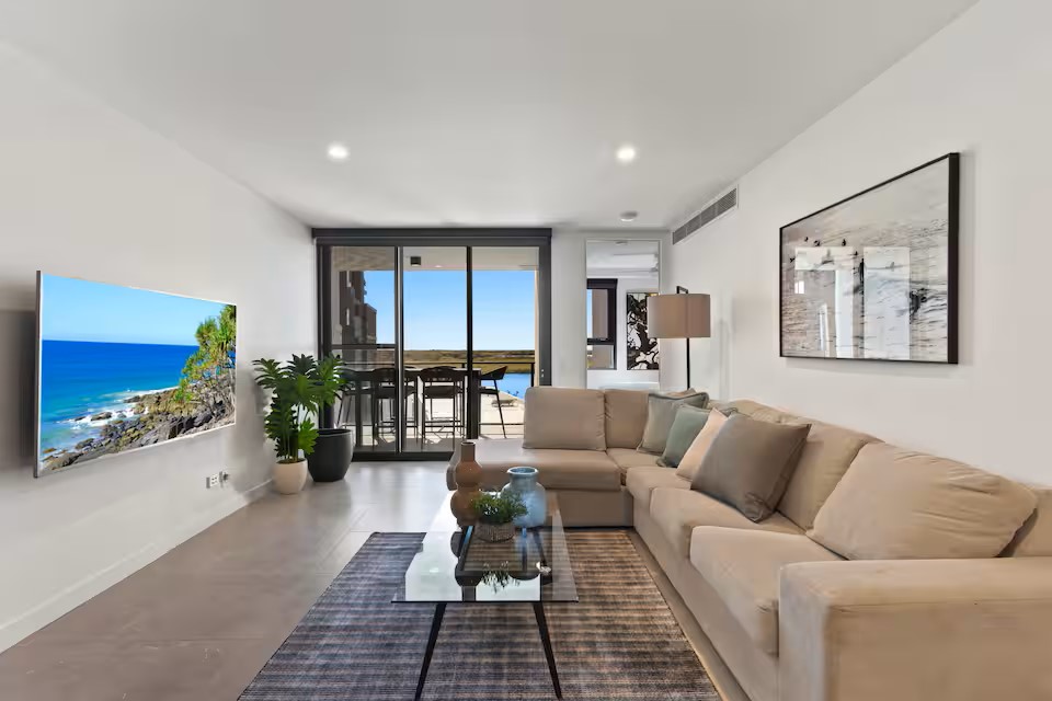 The living area features a plush, neutral-colored couch arranged around a modern glass coffee table. A large smart TV is mounted on the wall opposite a sliding door, which opens onto a balcony with scenic views. Natural light fills the room, complemented by indoor plants.