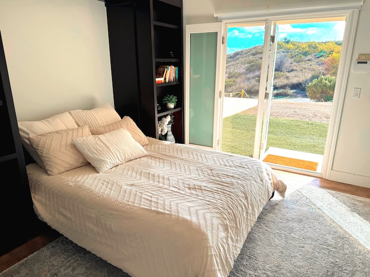 Chumba cha wageni cha ghorofa ya chini. Kitanda cha Malkia Murphy kwenye zulia la kijivu.    Mlango wa dirisha ulio na mlango tofauti wa kujitegemea ulio na mwonekano wa mazingira ya asili. Blinds za kuzima
 