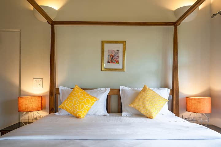 Drift off to sleep in this cosy bedroom at our Goa villa, featuring a charming four-poster bed, vibrant yellow accents, and warm lighting for a truly relaxing stay.